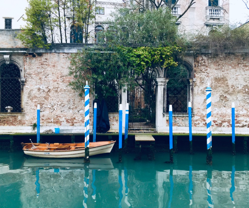 A small wooden boat and blue-and-white poles in a canal in front of a brick wall